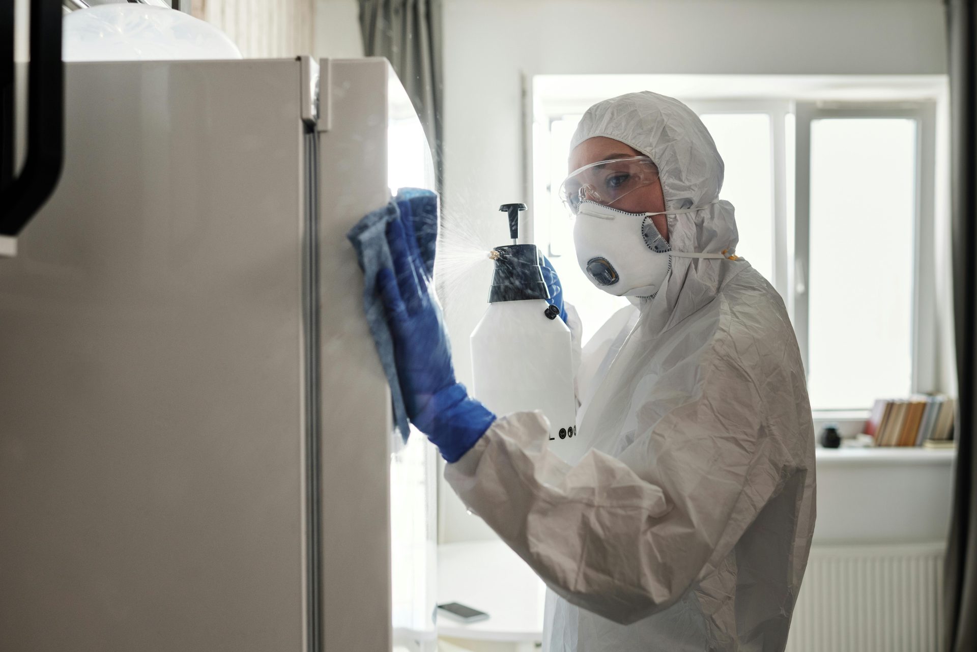 Person in protective gear sanitizing refrigerator at home for safety.