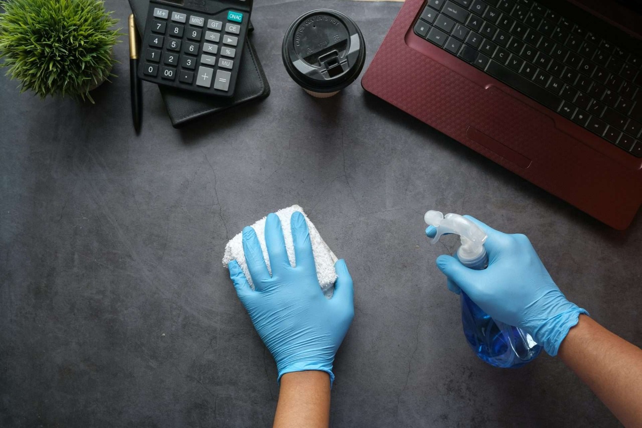 Gloved hands cleaning desk with disinfectant spray.